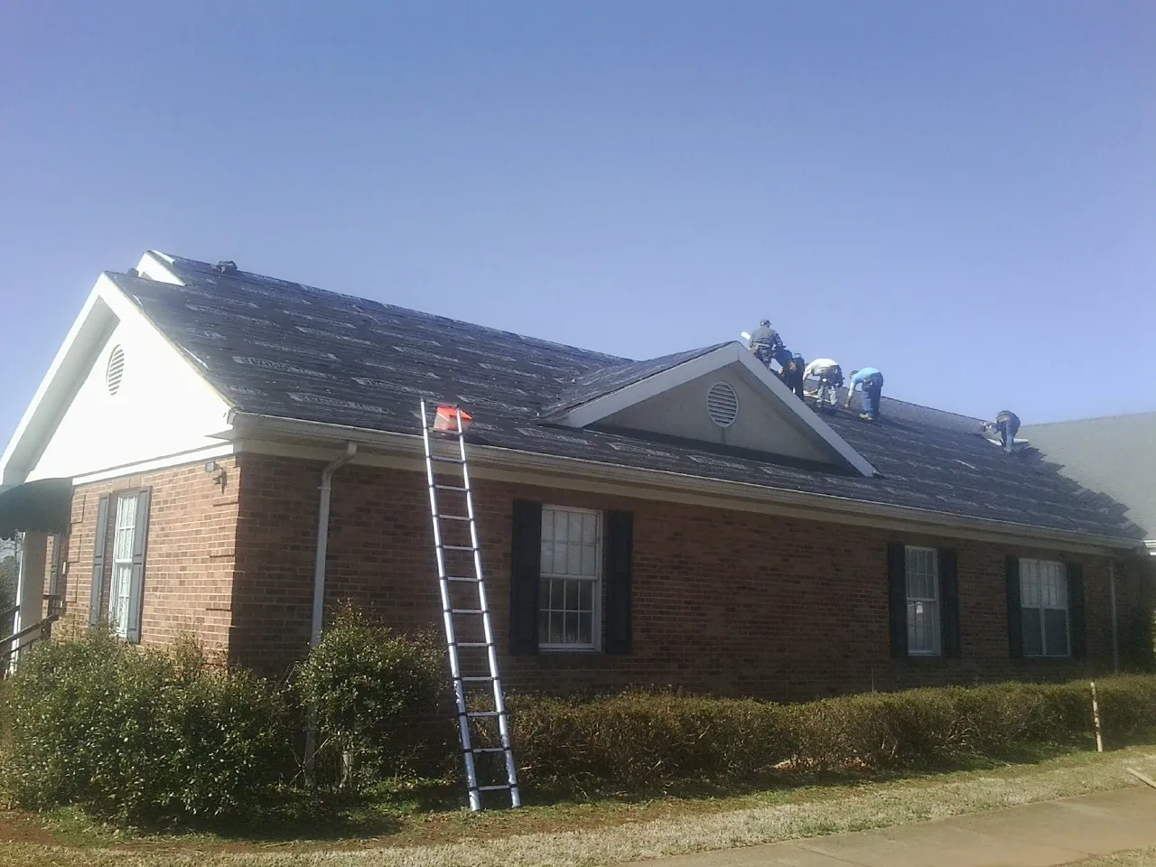 Crew tearing shingles off a brick ranch roof with a ladder set — Metal Roof Repair work by our crew in Vestal