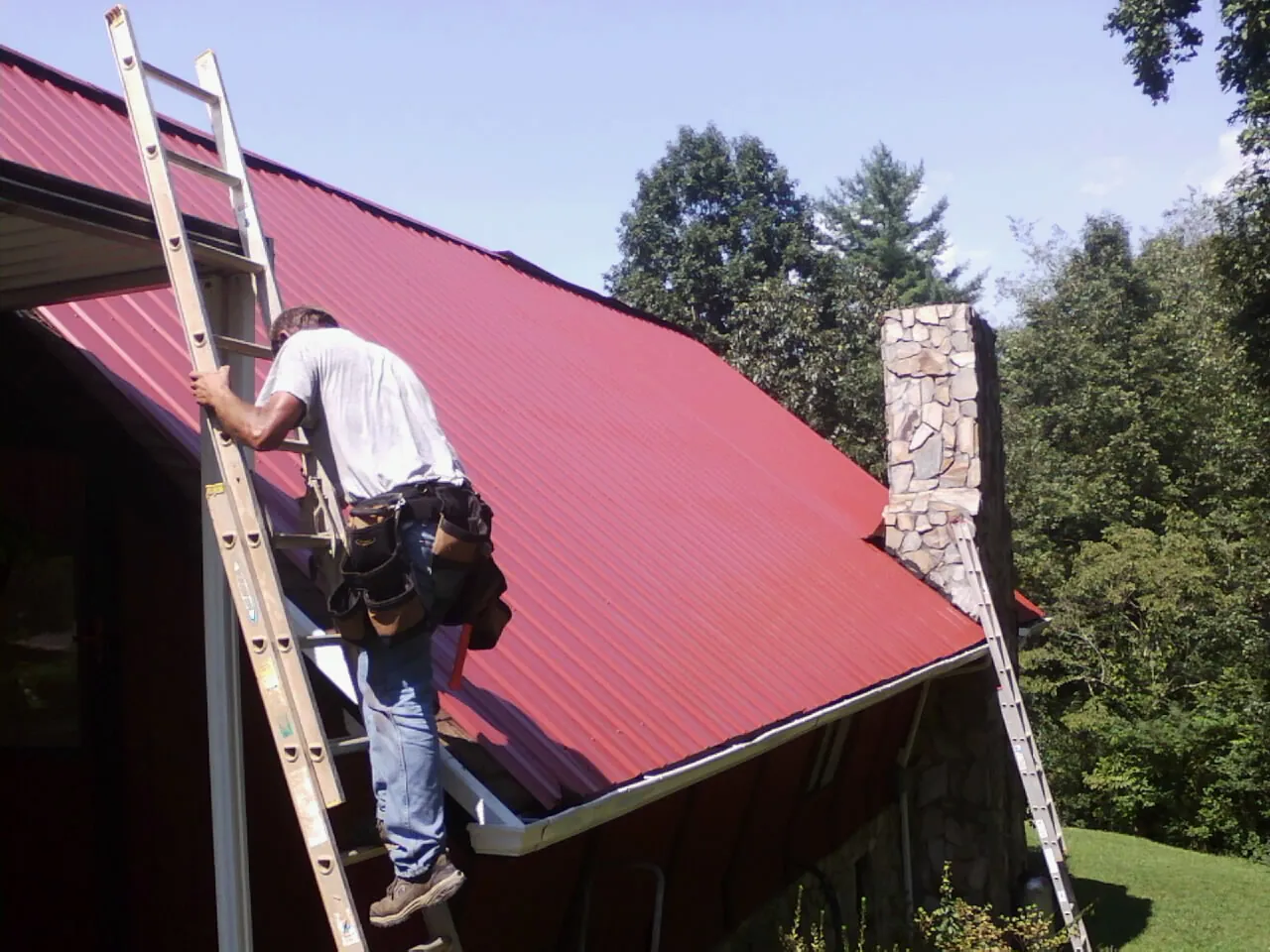Roofer on a ladder beside a red metal roof and stone chimney — Soffit & Fascia Repair by Roofing Brothers in Vestal